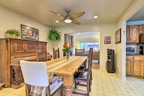 A dining area with a wooden table, chairs, and a ceiling fan. There is a refrigerator and a microwave in the kitchen area.