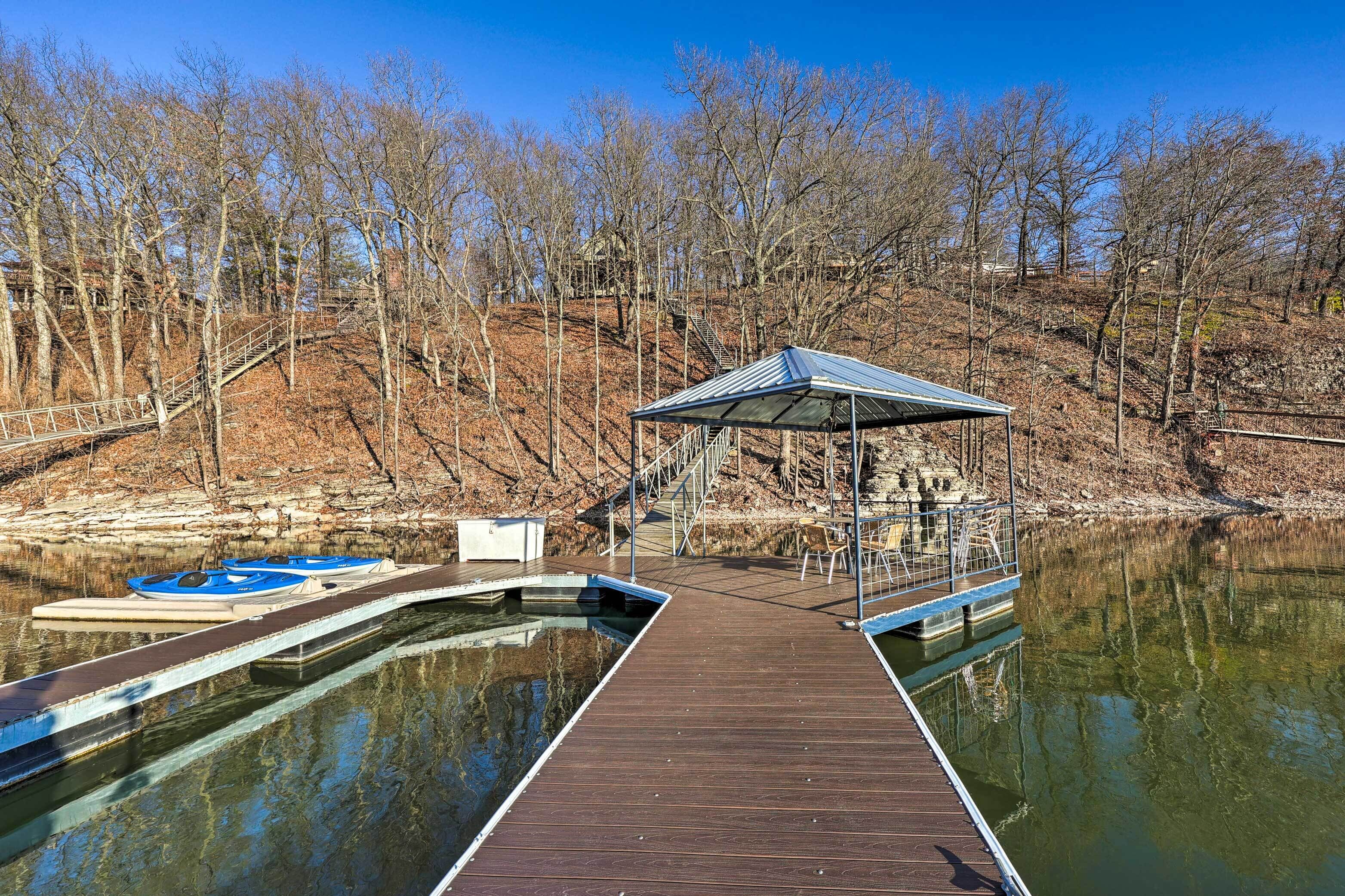 A wooden dock extending into a calm lake, with a covered seating area and a staircase leading up a hill.