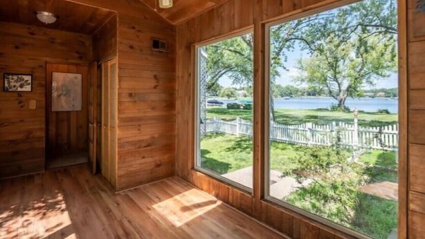 Une pièce de cabane en bois avec une grande fenêtre, un ventilateur de plafond et une porte donnant sur une autre pièce.