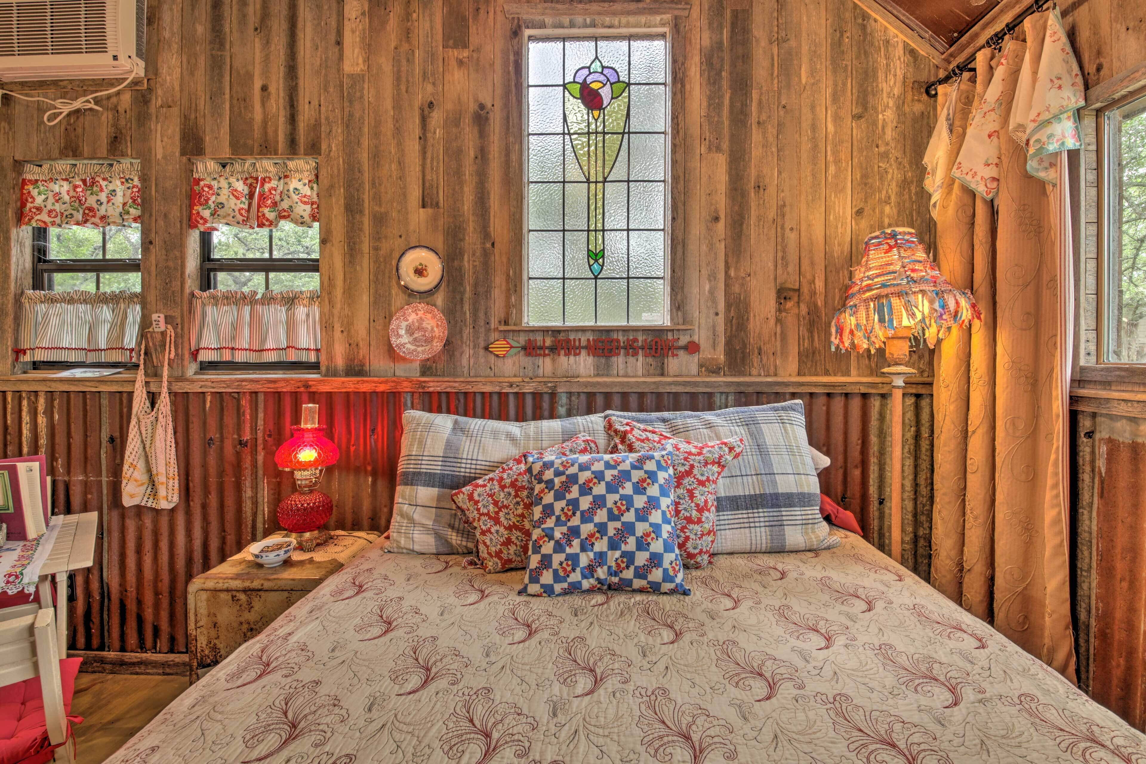 A rustic bedroom with wooden walls, a bed with patterned bedding, a stained glass window, and a red lamp.