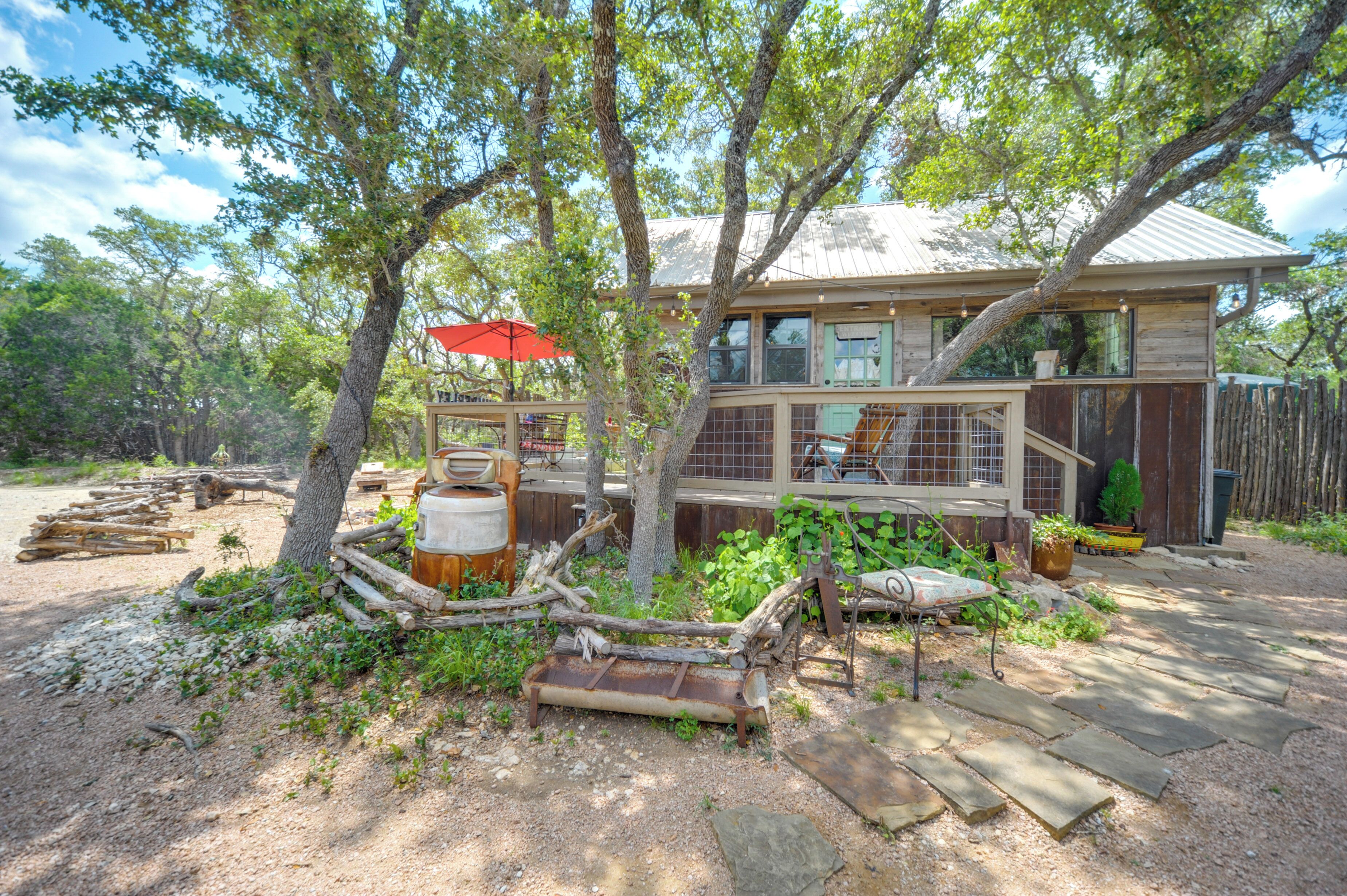 A rustic cabin with a wooden deck, surrounded by trees and a fenced yard.