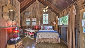 A rustic wooden room with a bed, a desk, and a chair. There is a ceiling fan, a window with curtains, and a wall-mounted air conditioner.