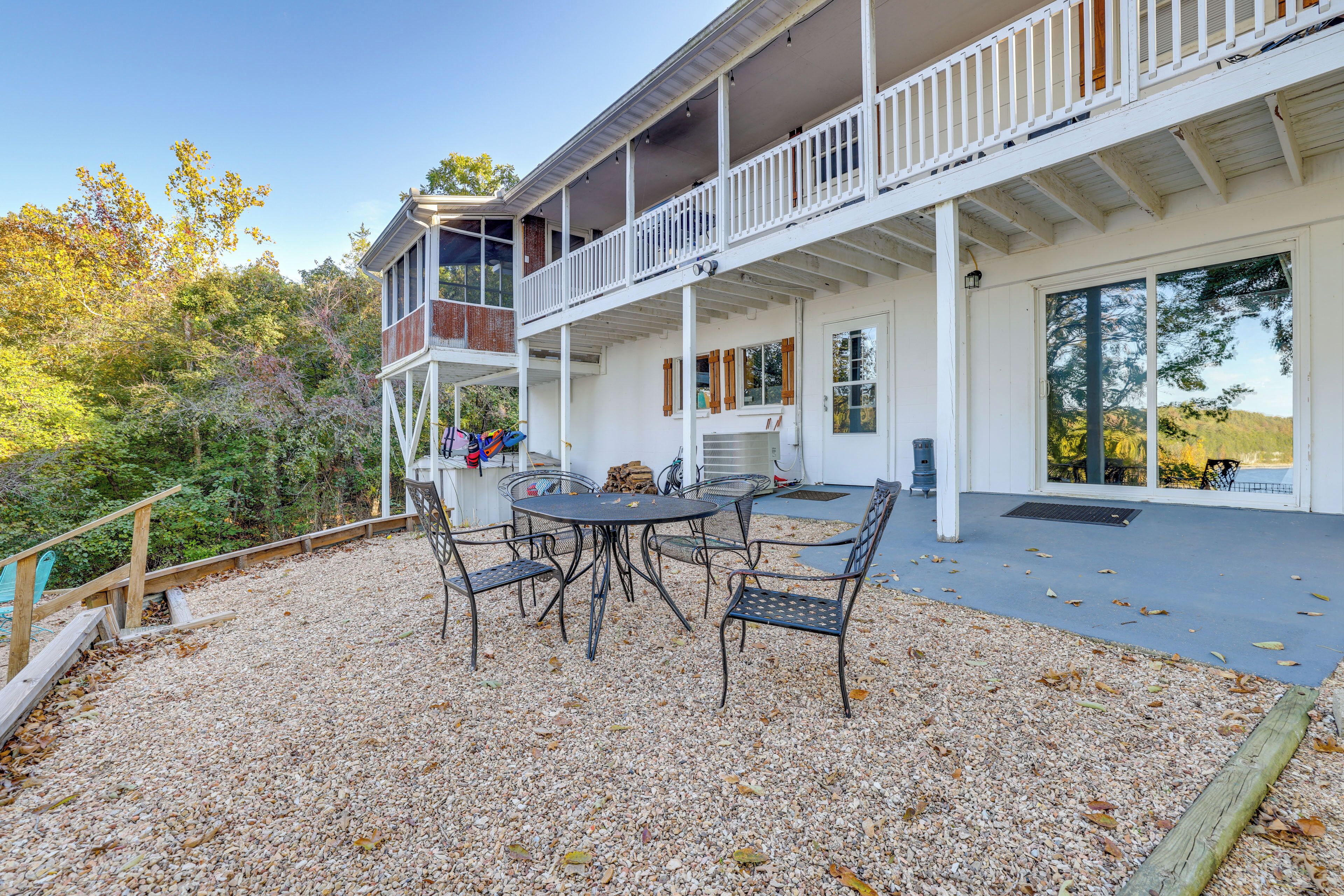 A two-story house with a balcony, a patio with a table and chairs, and a gravel ground.