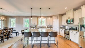 A modern kitchen with a central island, bar stools, and a dining area with a wooden table and chairs. - Lake Anna Home w/ Dock & Private Shoreline! (Bumpass)
