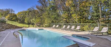 A swimming pool with a diving board, surrounded by lounge chairs and a fence.
