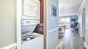 Een wasruimte met een witte wasmachine en droger, houten lambrisering, een eethoek met een tafel en stoelen, en uitzicht op het buitengebied.