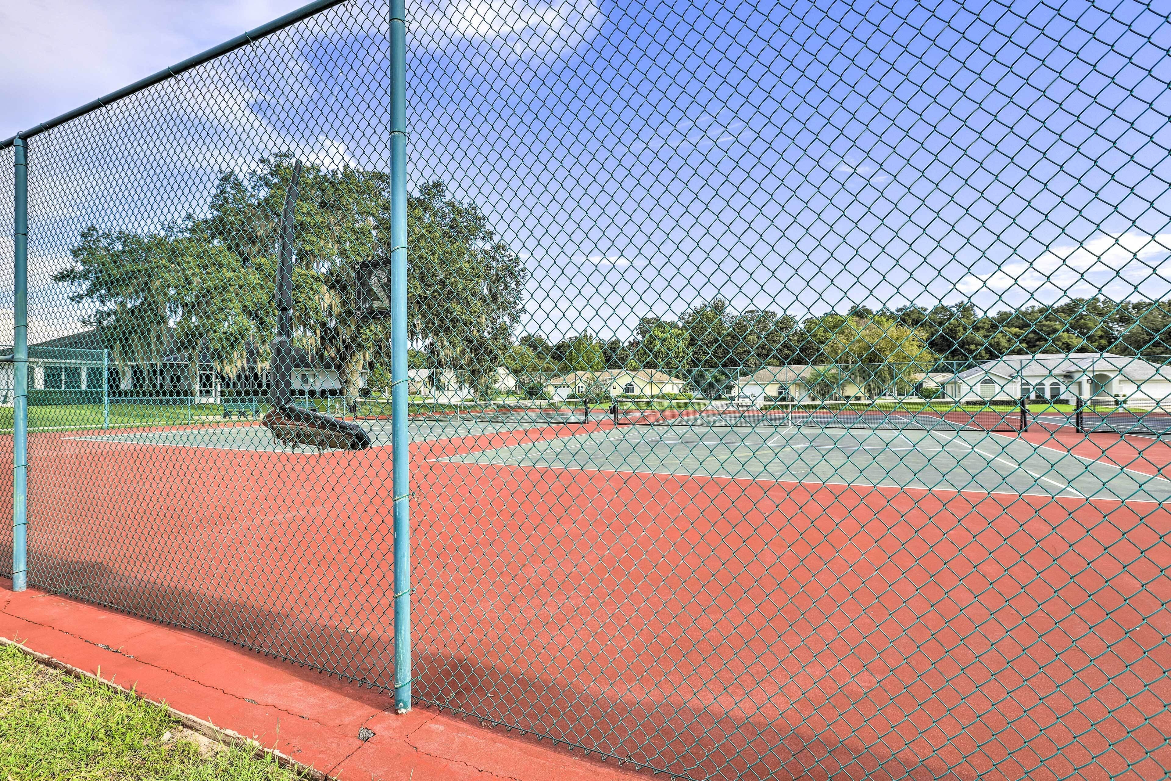 A fenced tennis court with a red surface, surrounded by a chain-link fence and trees in the background.