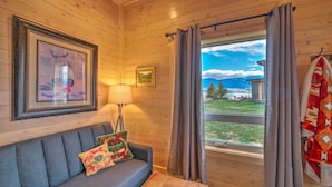 A cozy room with a wooden ceiling, a gray sofa with decorative pillows, a framed picture, a lamp, and a window with curtains. - Ennis Lake Studio w/ Kayak, Pool & Fire Pit! (McAllister)