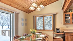A dining area with a wooden table set for six, a chandelier, and a view of a snowy landscape through large windows. - Cedar Cottage, Private Beach on Lake Leelanau (Cedar)