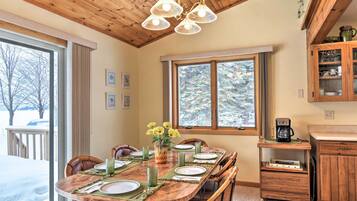 A dining area with a wooden table set for six, a chandelier, and a view of a snowy landscape through large windows.