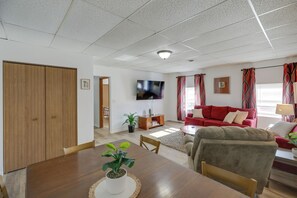 A living room with a wooden dining table, a red sofa, and a television mounted on the wall. - Bright Yankton Apartment Near Meridian Bridge (Yankton)