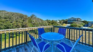 A wooden deck with a round table and blue chairs, overlooking a beach and a house.