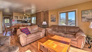 A spacious living area with a large brown leather sofa, a wooden coffee table, and a dining table with chairs. A brick fireplace and a large window with a view of buildings are visible.