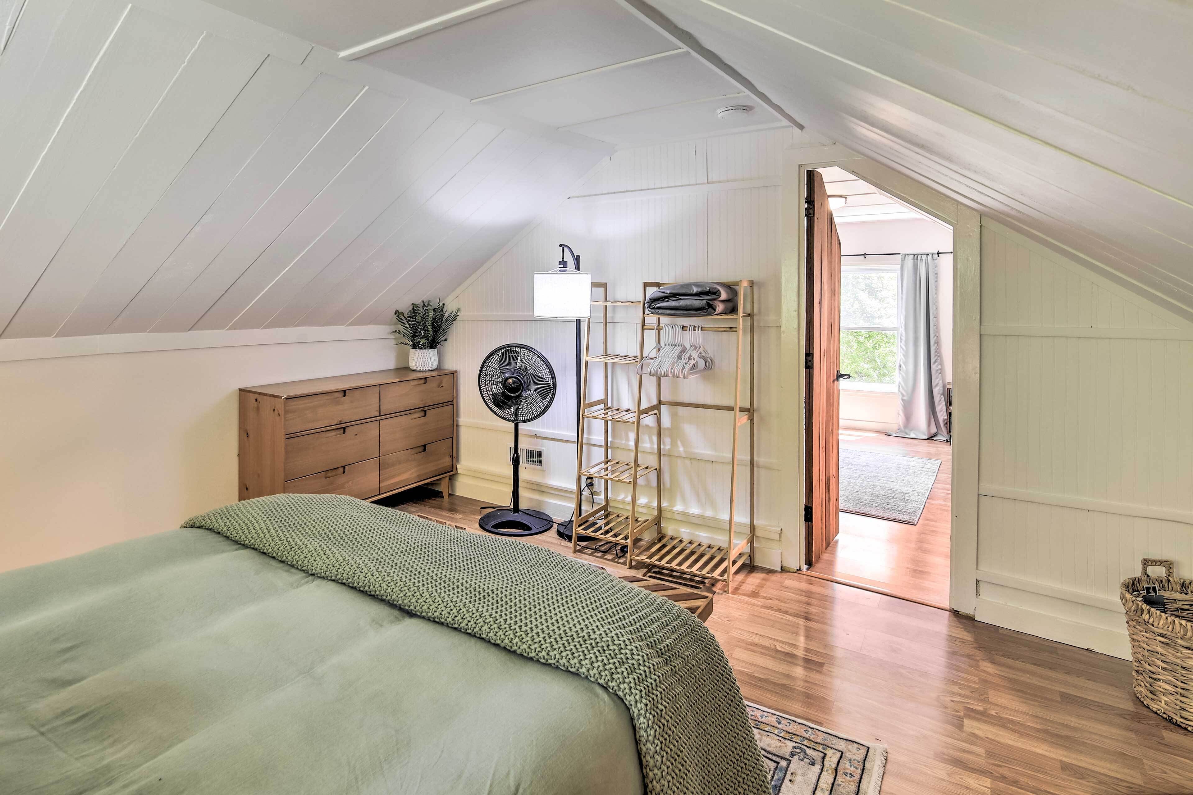 Quarto com teto inclinado, cômoda de madeira, cama com cobertor verde, ventilador e um varal.
