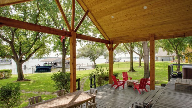 A covered outdoor deck with wooden furniture, a view of a lake, and surrounding greenery. - Stunning Lakefront Home: Hot Tub & Game Room! (Mabank)