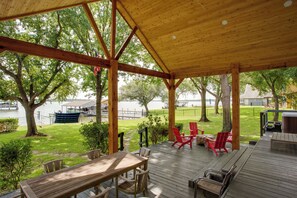 A covered outdoor deck with wooden furniture, a view of a lake, and surrounding greenery.