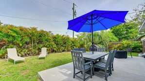 An outdoor patio with a table and chairs, a blue umbrella, and lush greenery.