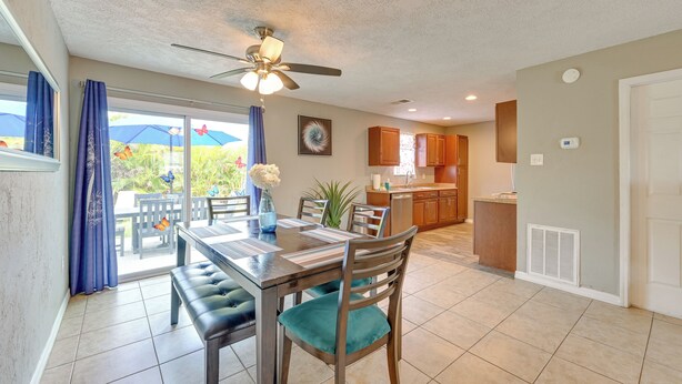 A dining area with a table, chairs, and a ceiling fan. There is a kitchen in the background with wooden cabinets and a window.