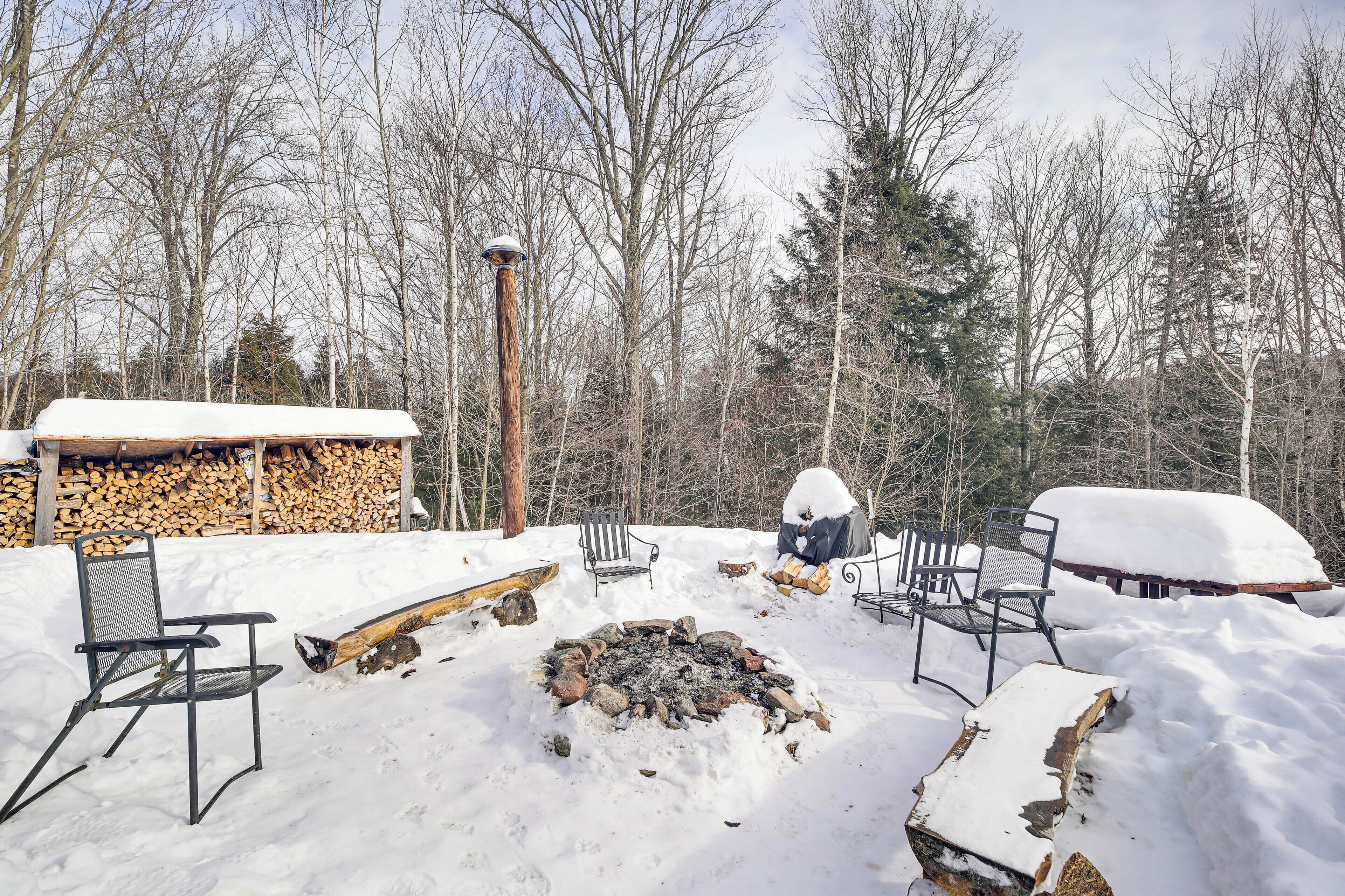 A snow-covered outdoor seating area with a fire pit, surrounded by trees and a stack of firewood.