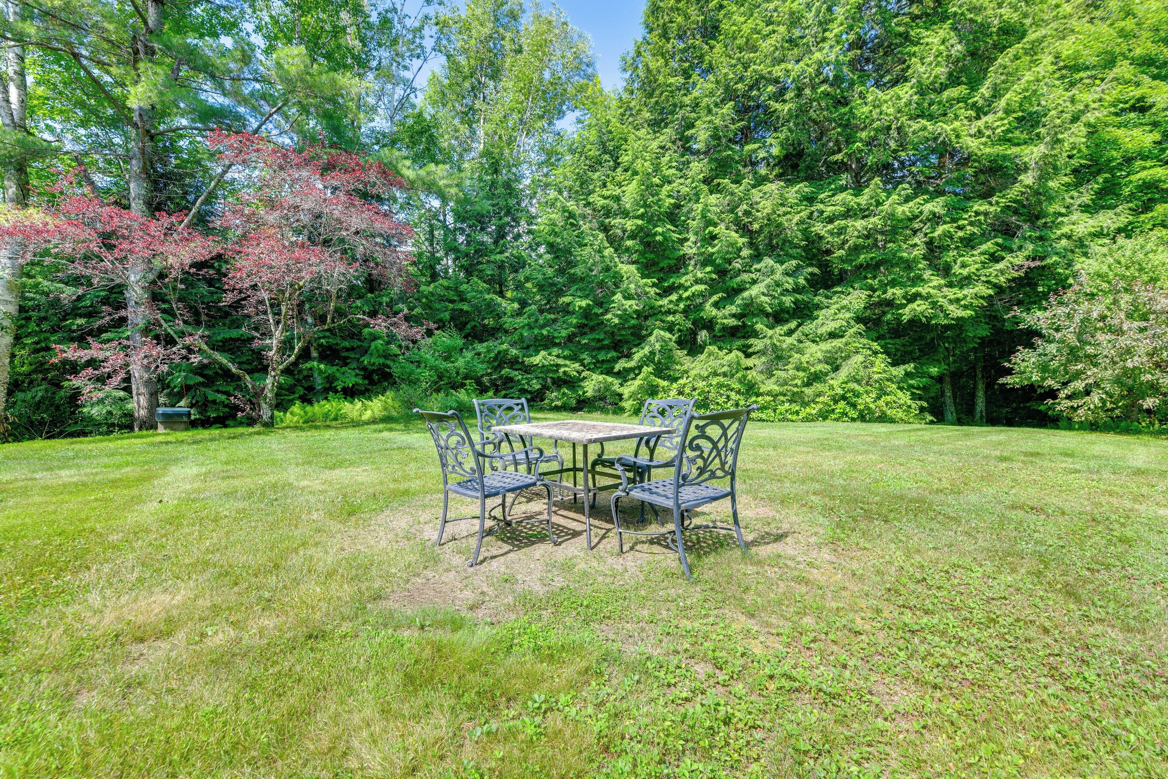 A grassy outdoor area with a metal table and chairs, surrounded by trees and greenery.