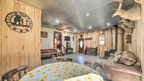 A rustic interior with wooden walls, a round table with a sunflower tablecloth, a leather sofa, and mounted deer heads. - Charming New Concord Cabin on 50-acre Farm! (New Concord)