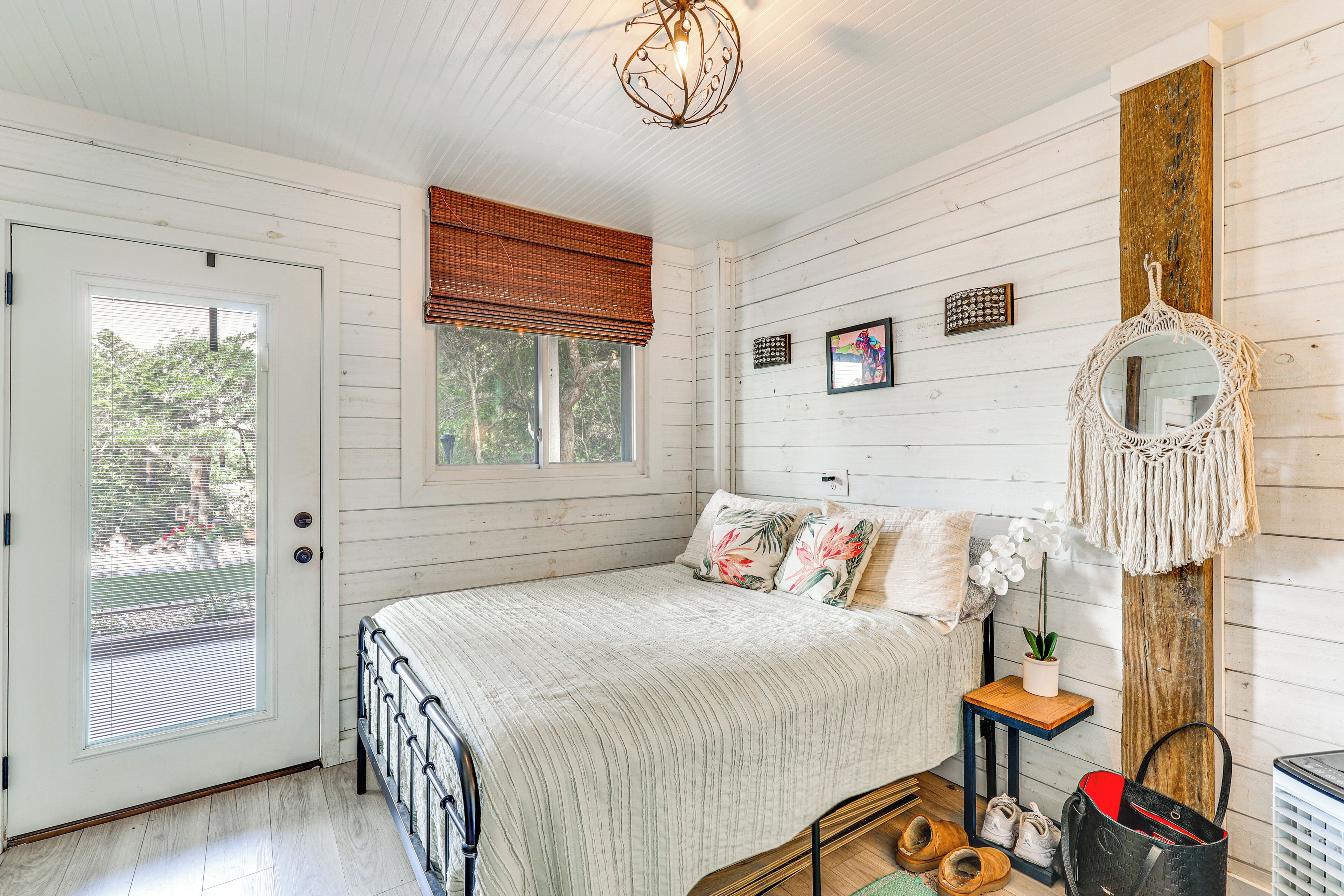 A bedroom with a bed, a door, a window, a wall-mounted mirror, and a small table with a plant.