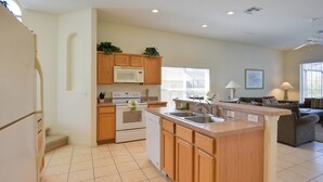 A modern kitchen with wooden cabinets, a built-in microwave, and a dishwasher. There is a large island with a sink and a window above the sink. The kitchen is adjacent to a living area with a couch and a lamp. - Family Retreat w/ Pool: 10 Mi to Disney! (Clermont)