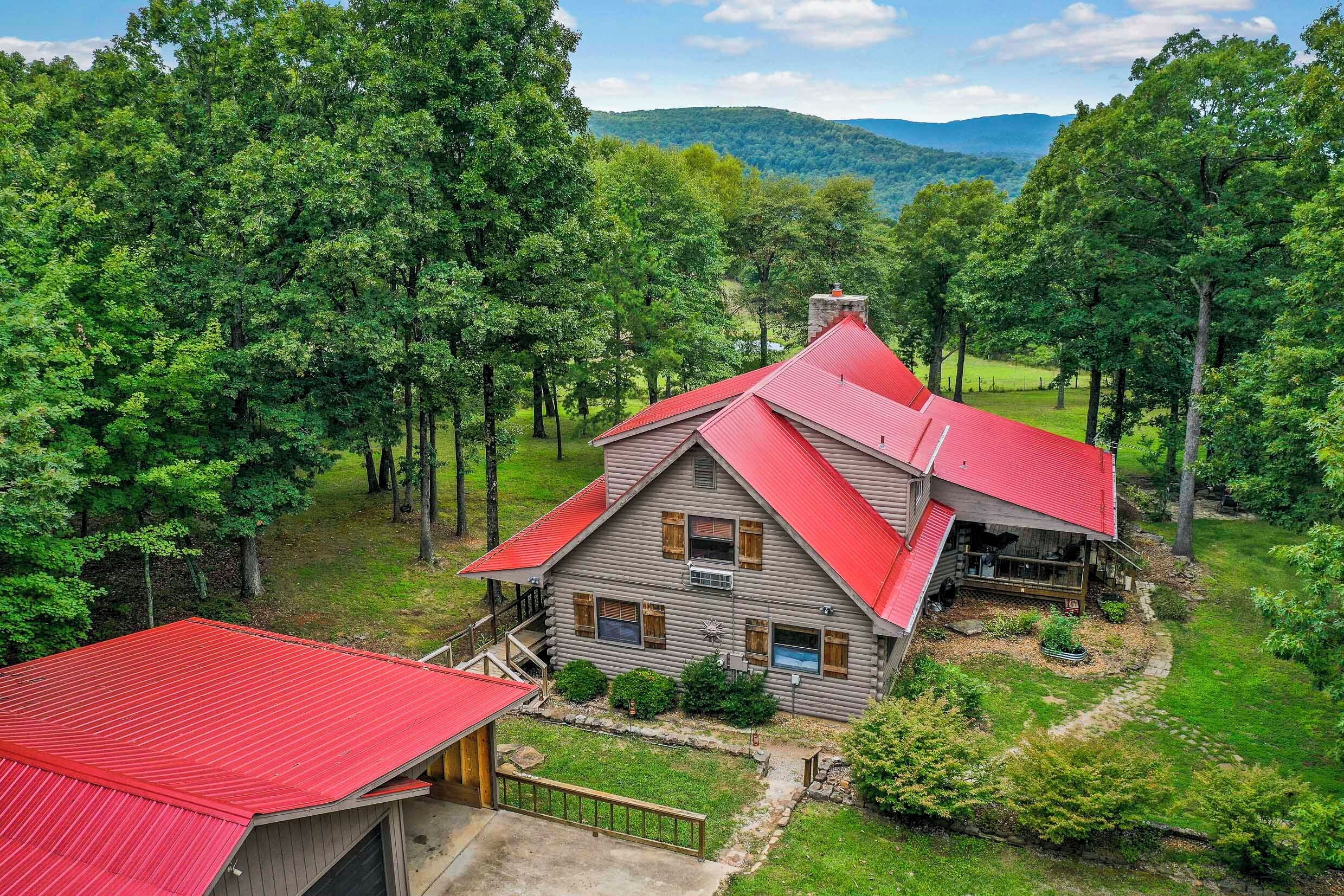 A log cabin with a red roof, surrounded by lush greenery and trees, with a view of distant mountains.