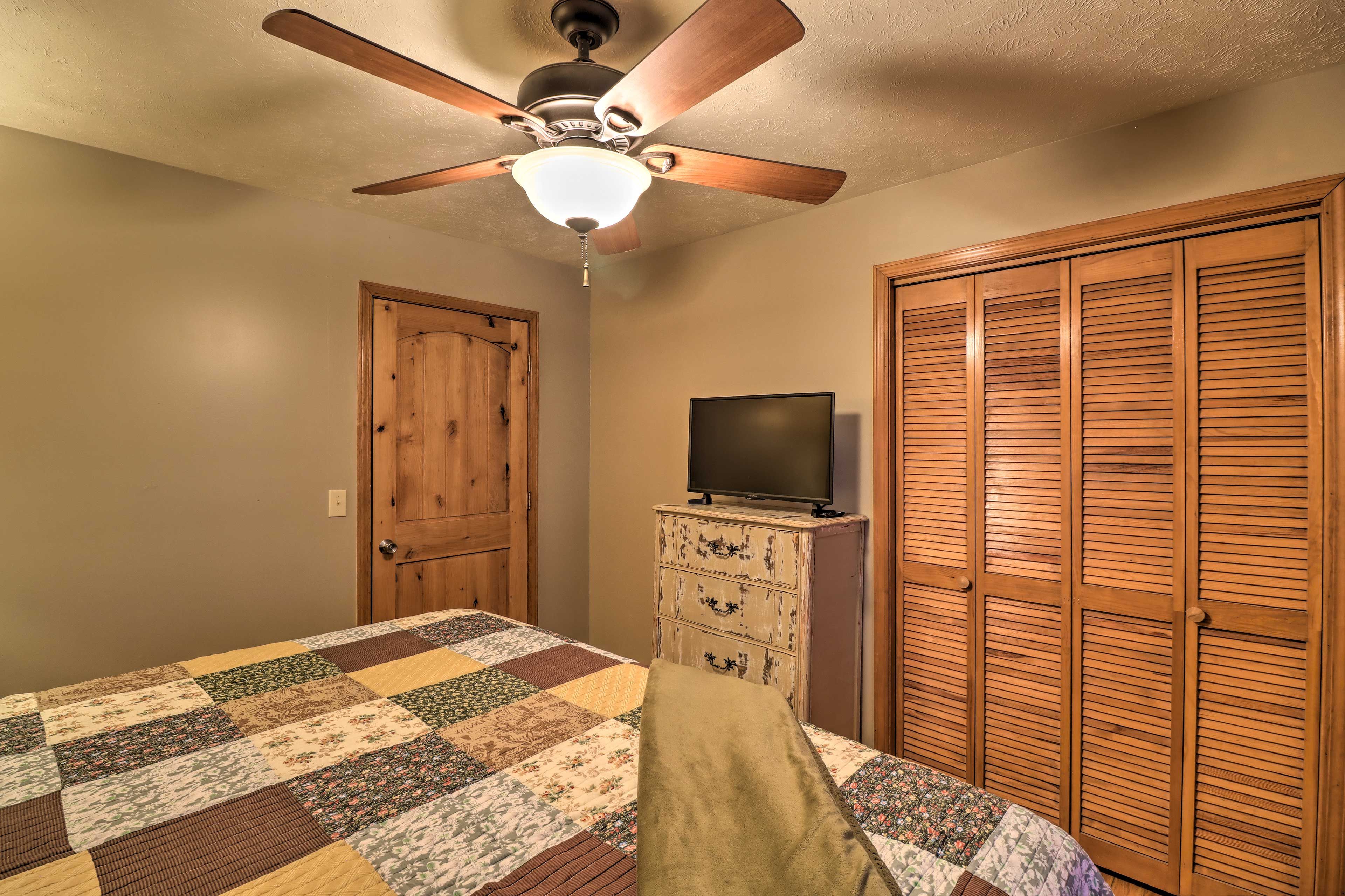 A bedroom with a quilted bedspread, a wooden dresser, a television on top, and a ceiling fan.