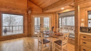 A wooden interior with a dining table set for four, a kitchen area with a microwave, and large windows offering a view of the outdoors.