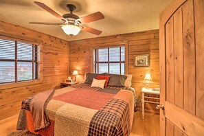 Un dormitorio de cabaña de madera con una cama, dos mesitas de noche, un ventilador de techo y una ventana con persianas.