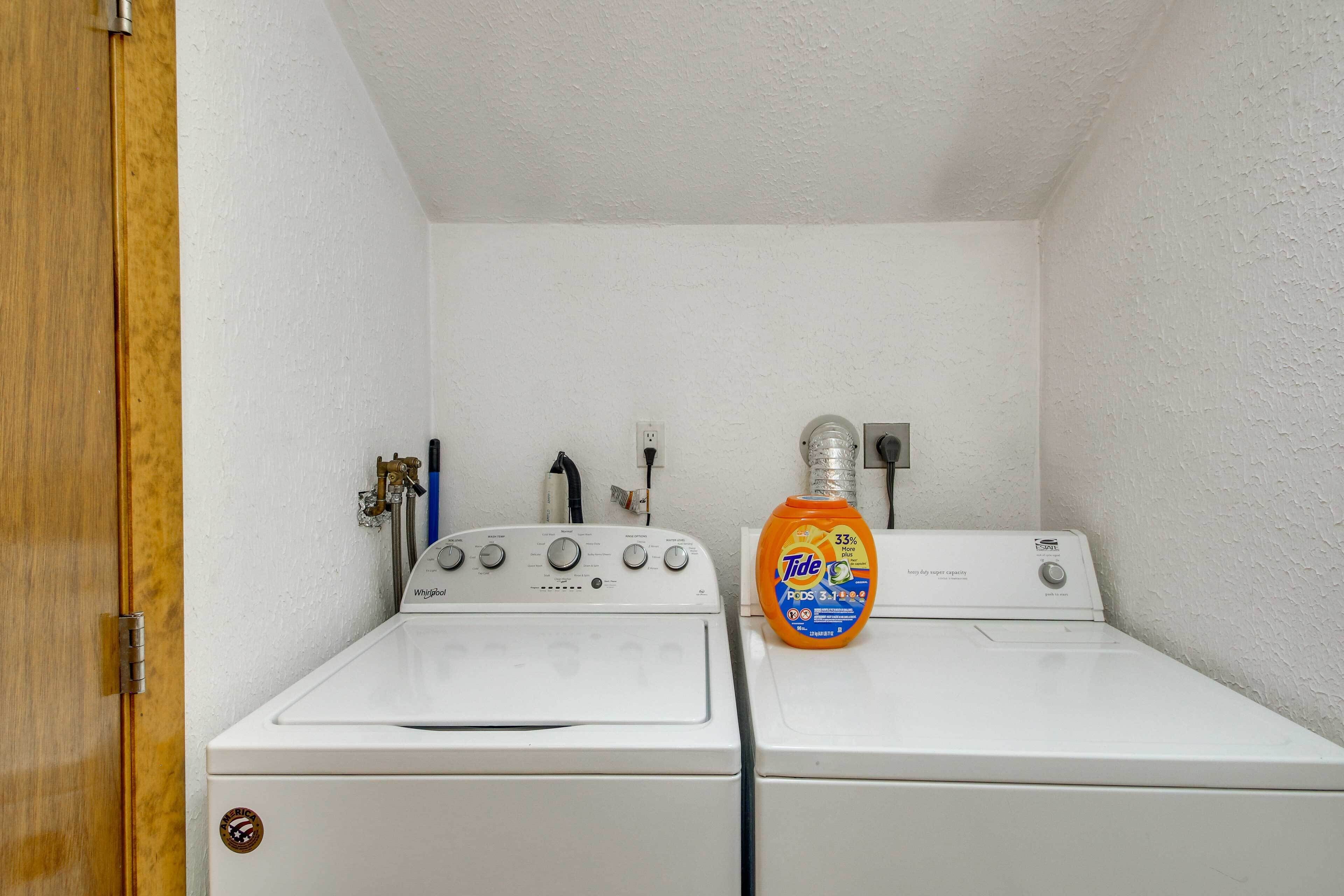 A laundry room with two white front-loading washers and dryers, a Tide detergent bottle, and a water heater.