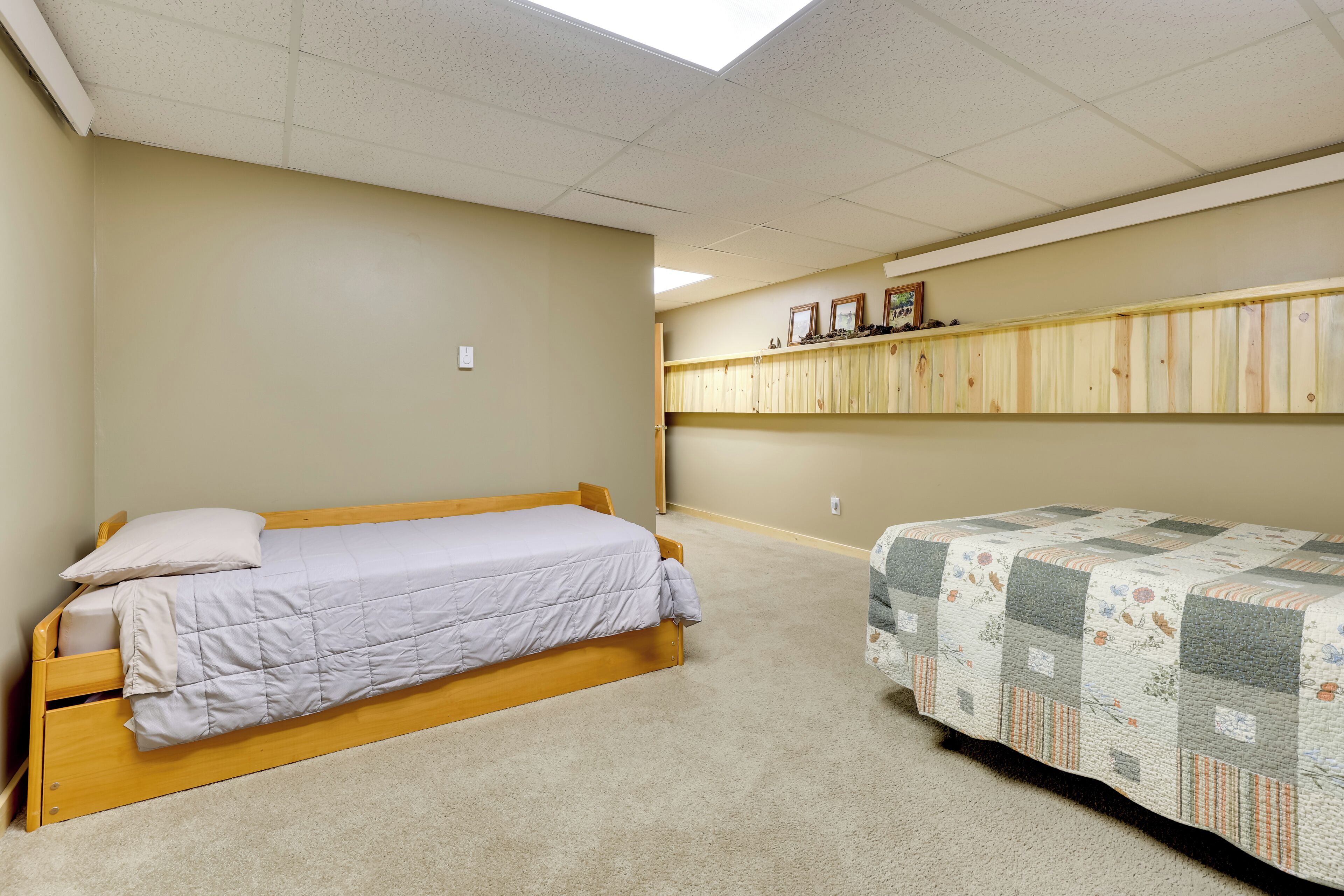 A bedroom with two beds, a ceiling fan, a light switch, and a wall-mounted shelf with framed pictures.