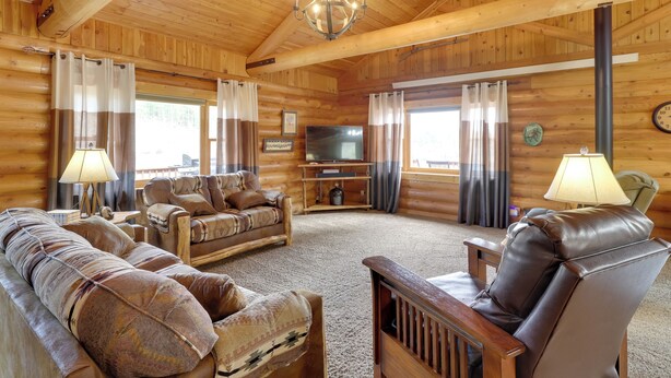 A log cabin interior with a wooden ceiling, a chandelier, a television, leather sofas, and a fireplace.