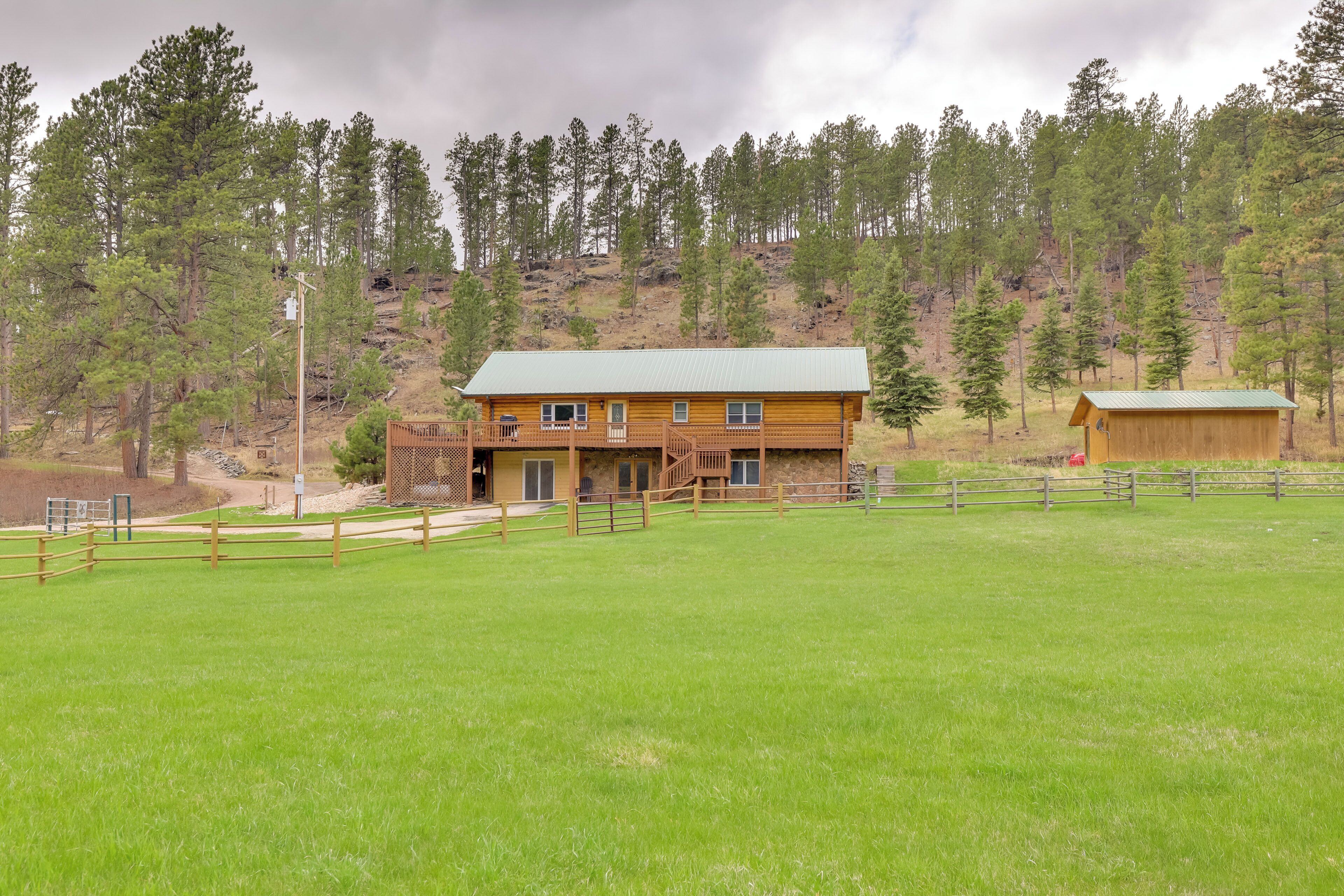 A wooden cabin with a green roof, surrounded by a fenced yard and a forested hillside.