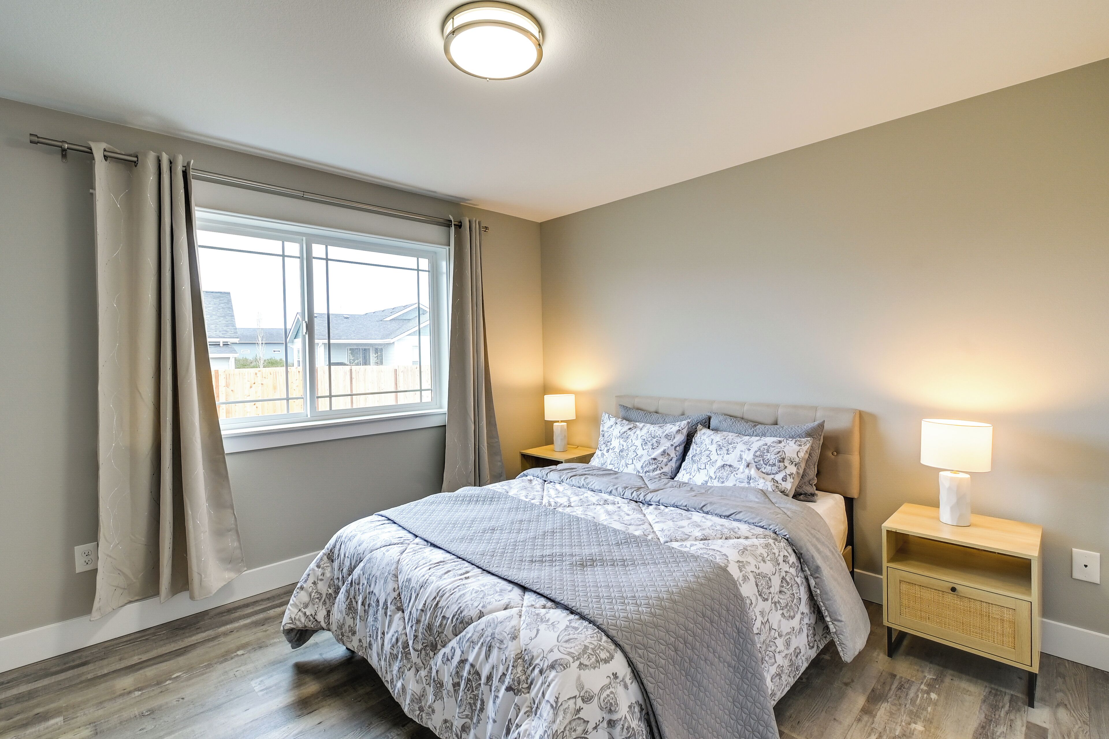 A bedroom with a large bed, two bedside tables with lamps, a window with curtains, and a view of buildings outside.