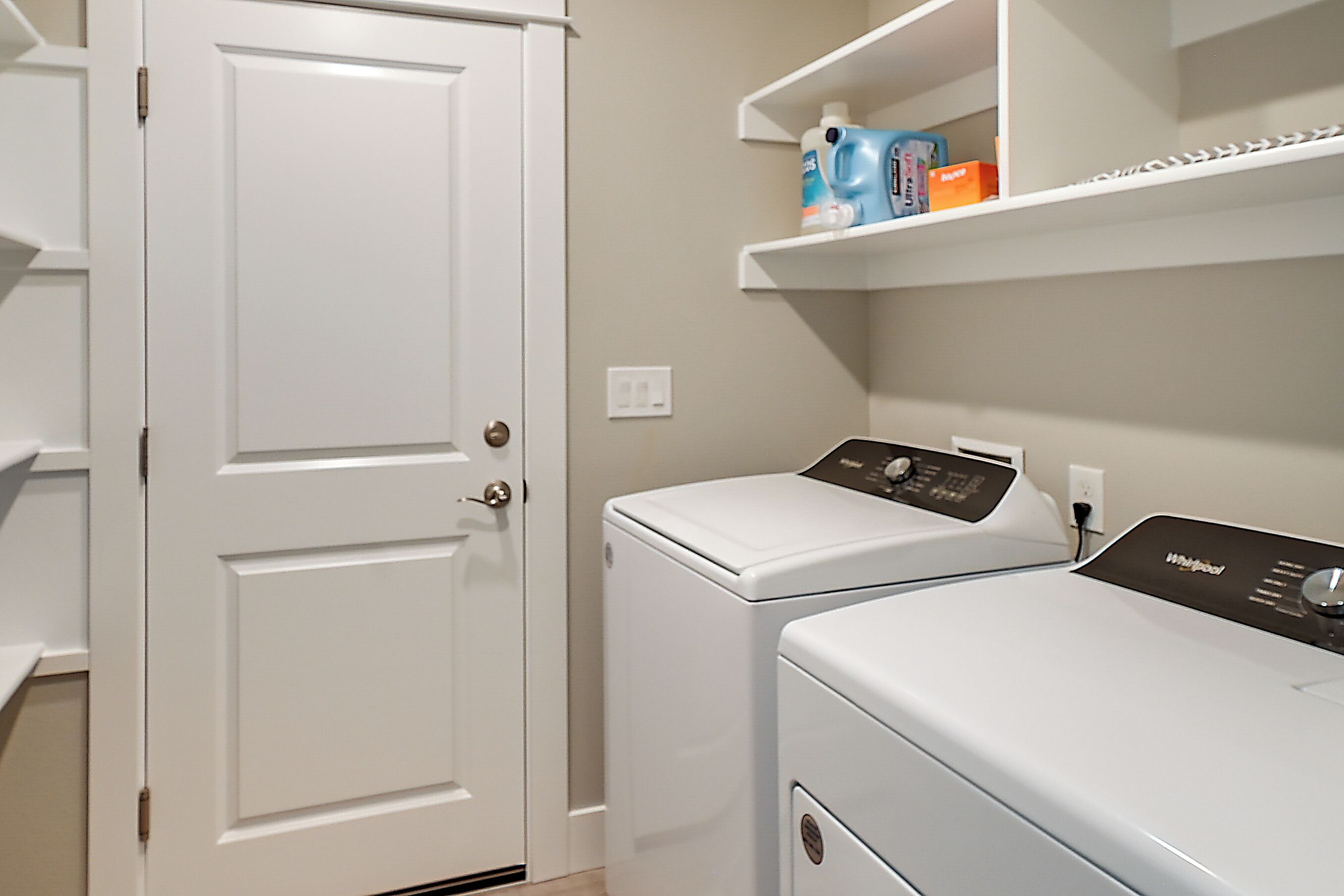 A laundry room with two white front-loading washers, a white door, and shelves above holding laundry supplies.