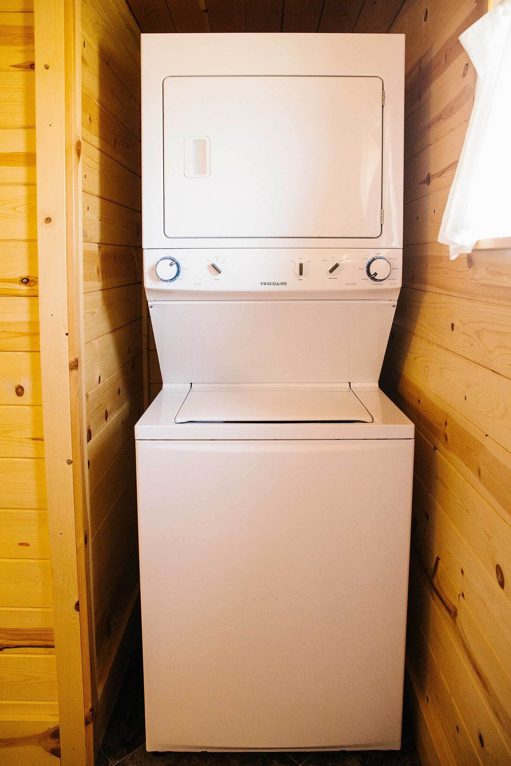 Een witte wasmachine in een houten blokhut, met een houten wand en een witte handdoek aan de rechterkant.