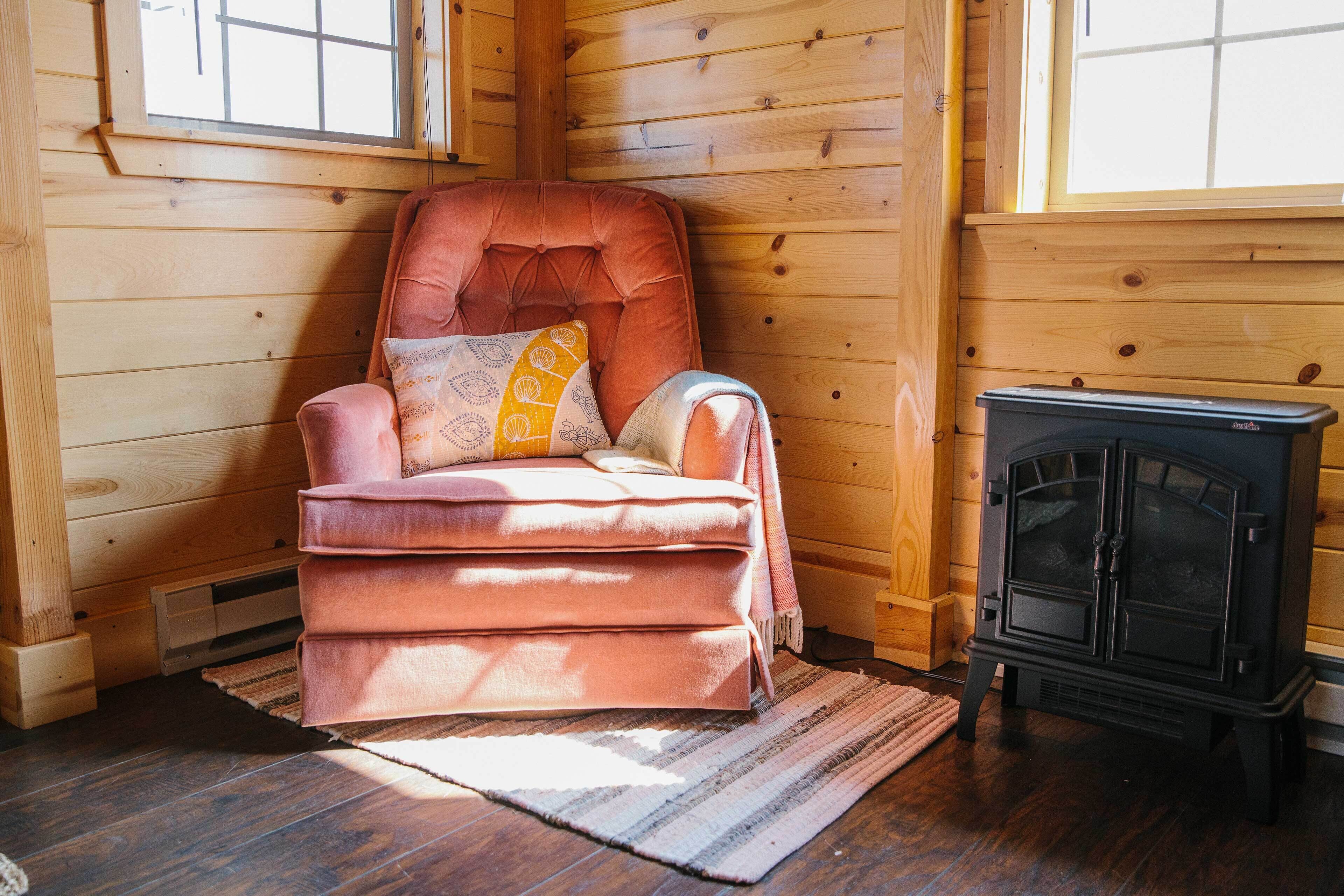 Um quarto de cabana de madeira aconchegante, com uma poltrona rosa macia, uma lareira a lenha preta e paredes de madeira.