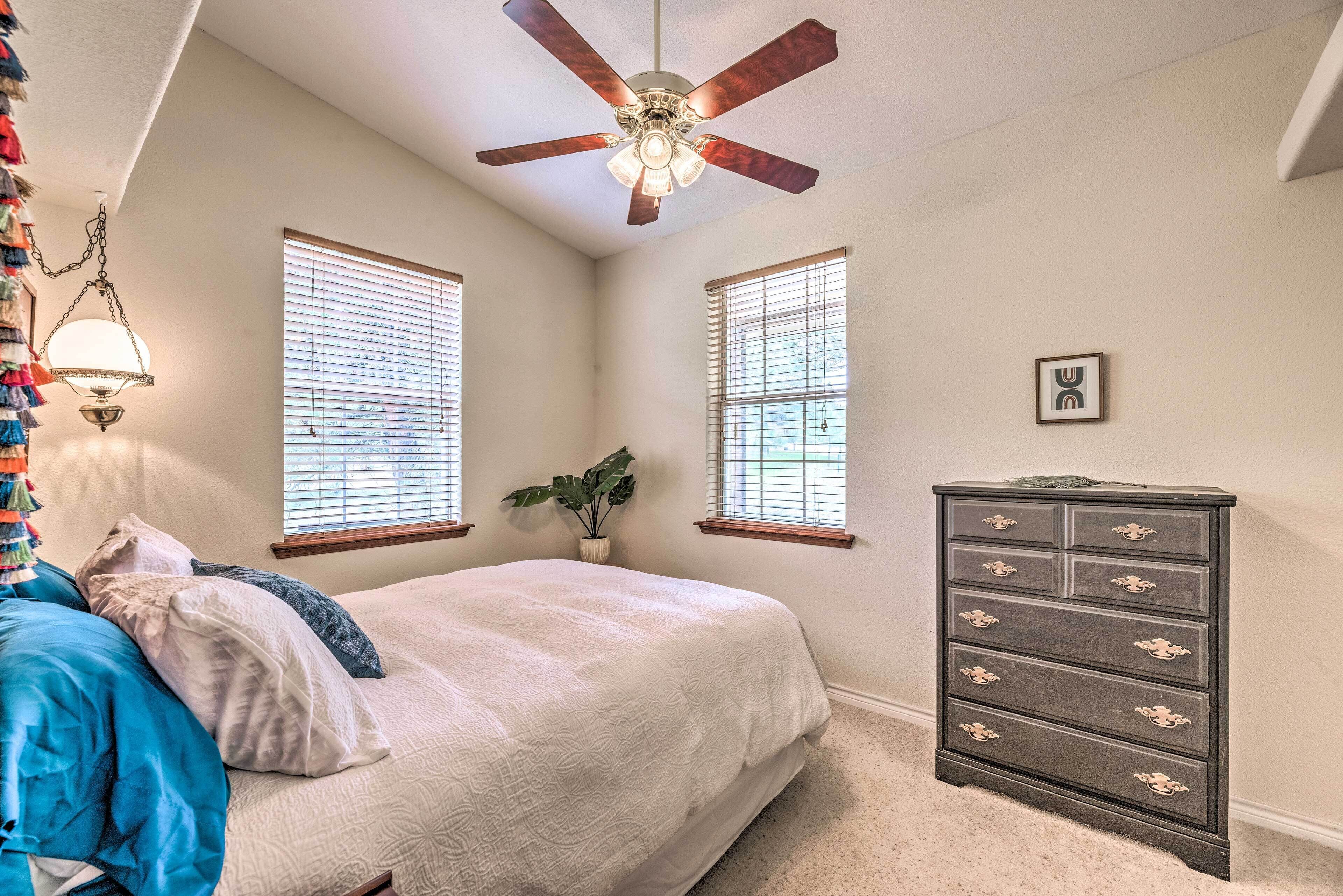 A bedroom with a bed, dresser, ceiling fan, windows, and a plant.