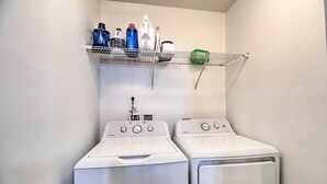 Un área de lavandería con dos lavadoras y secadoras frontales blancas, un estante metálico superior con varias botellas y recipientes, y una pared blanca.