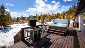 Een houten terras met een jacuzzi en een barbecue, met uitzicht op een besneeuwd landschap met bomen en een berg op de achtergrond.
