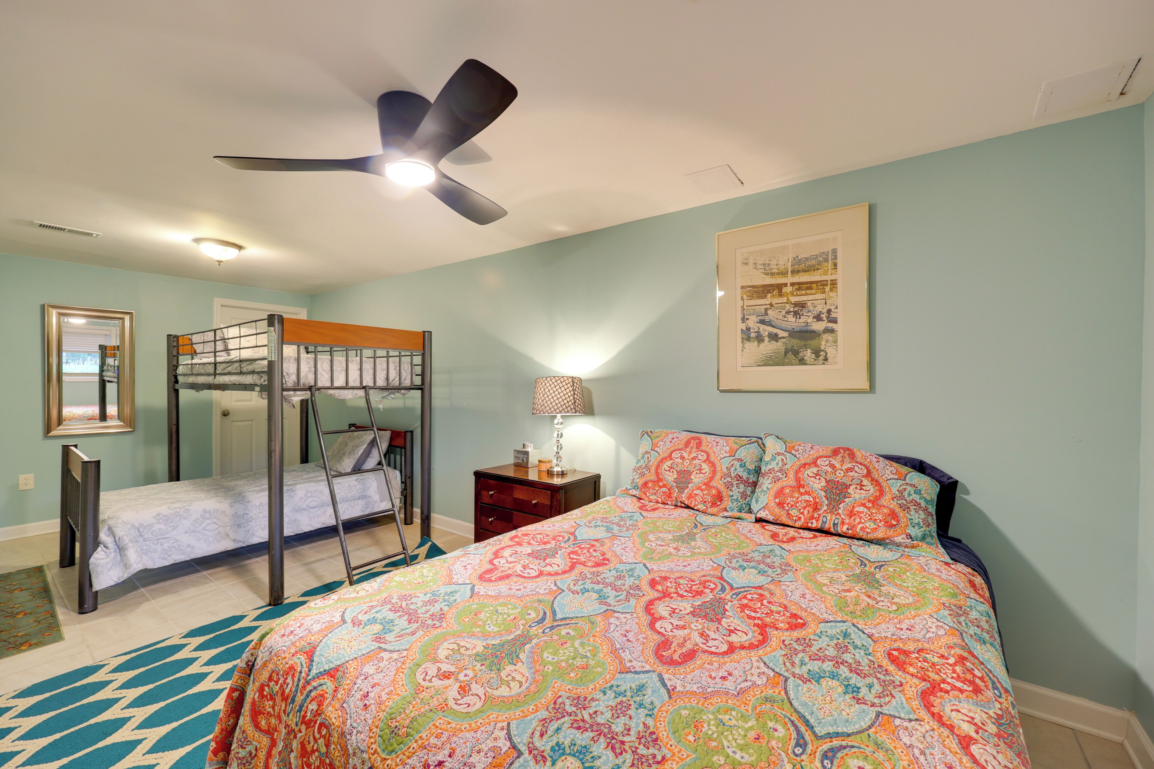 A bedroom with bunk beds, a bed with patterned bedding, a ceiling fan, a lamp, and a framed picture on the wall.