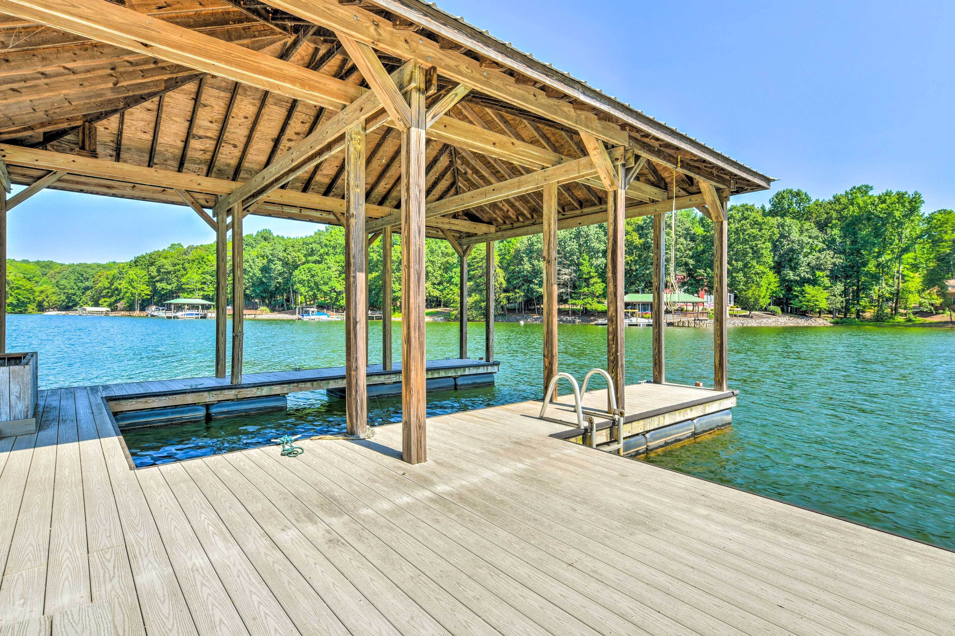 A wooden dock with a covered area and a pool, overlooking a lake with trees and buildings in the background.