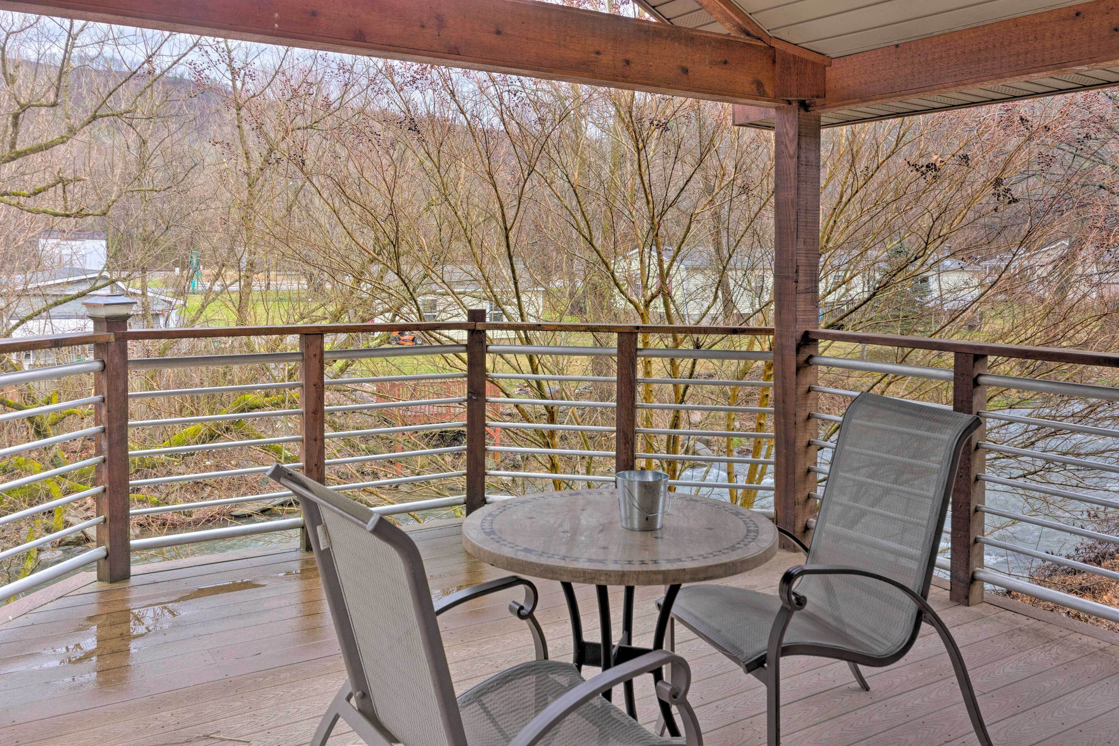 A covered outdoor area with a round table and two chairs, a metal railing, and a view of trees and buildings in the background.