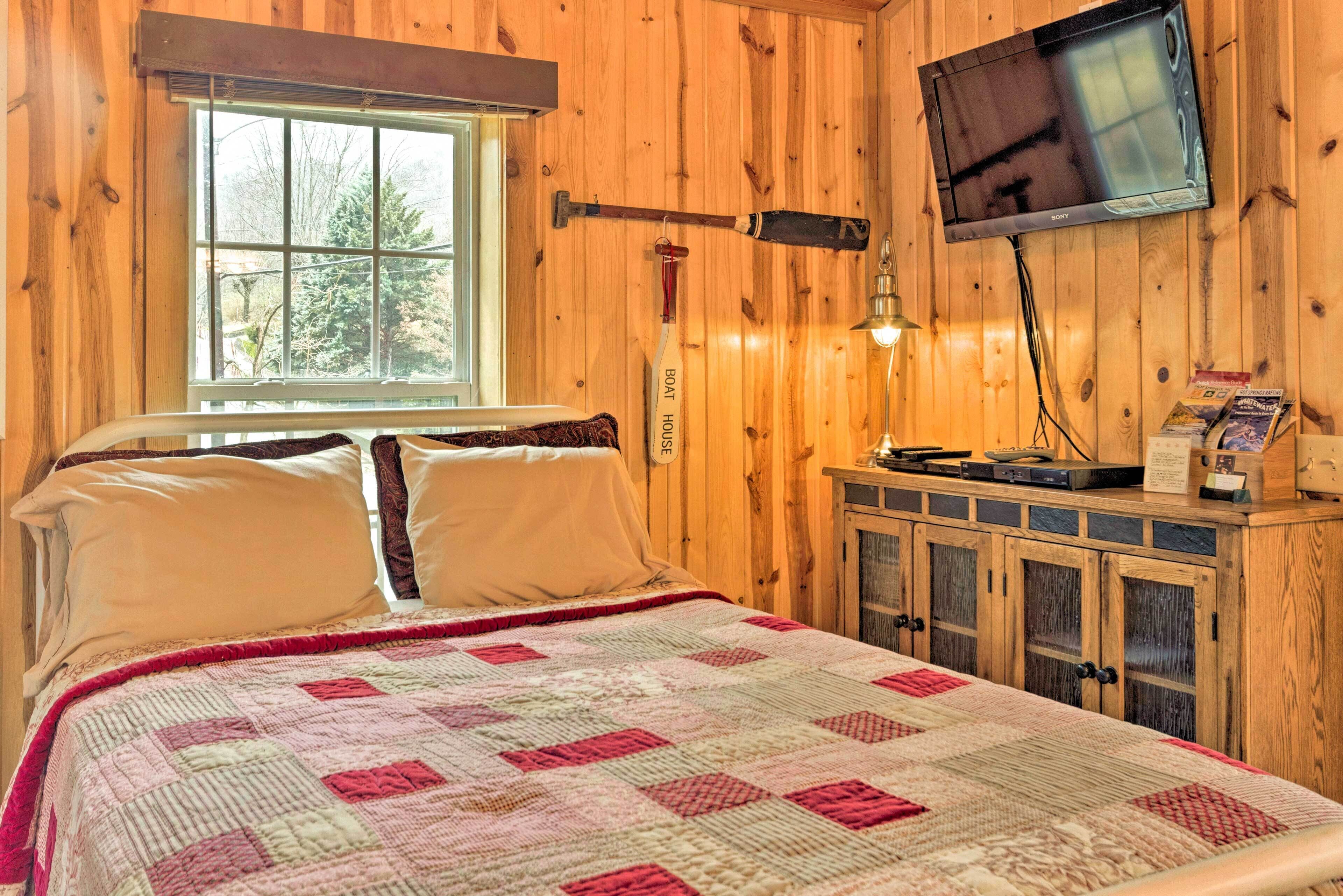 A wooden cabin room with a bed, a television mounted on the wall, and a window with a view of trees.