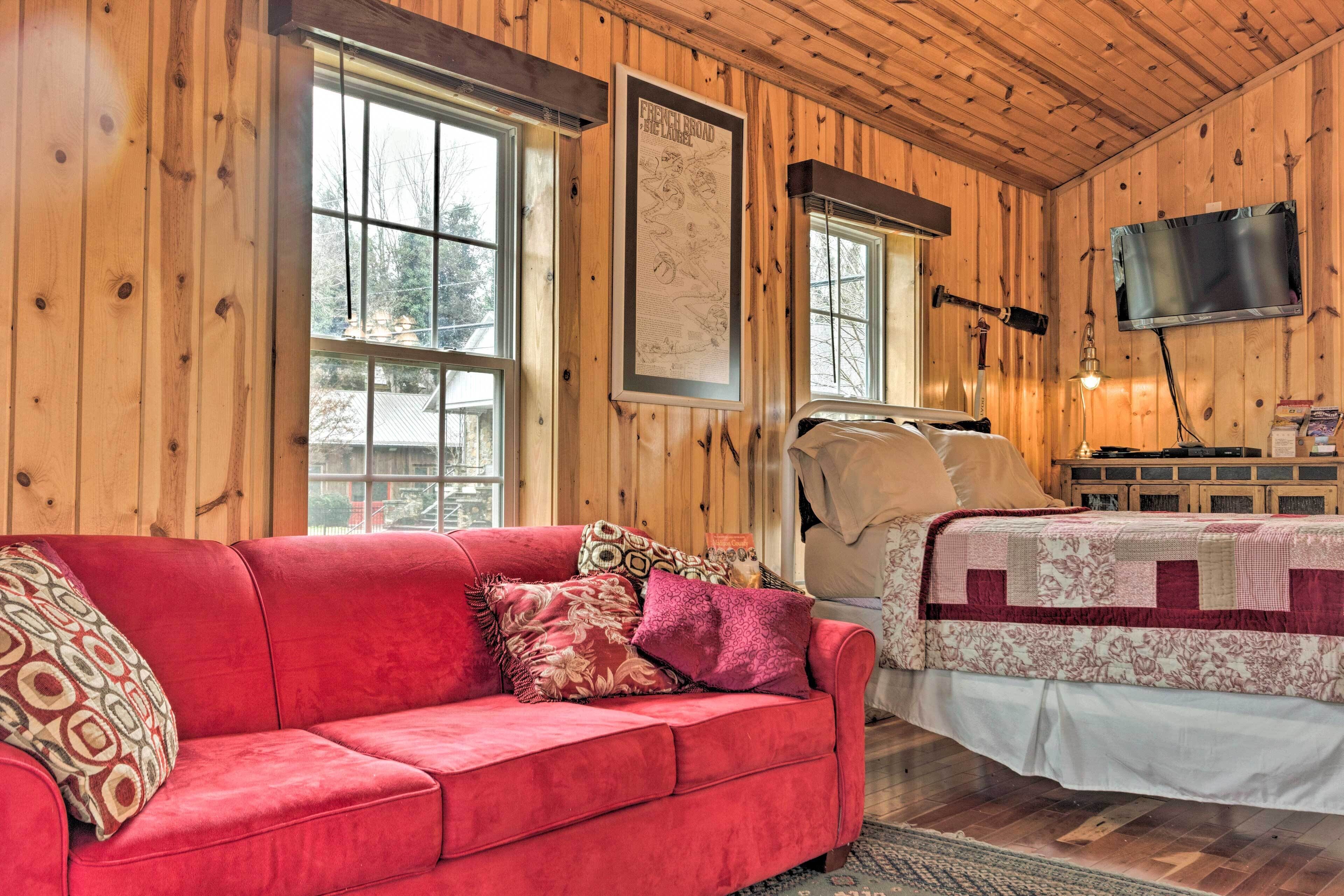 A cozy room with a red sofa, a bed with a floral quilt, wooden walls, and a mounted TV.