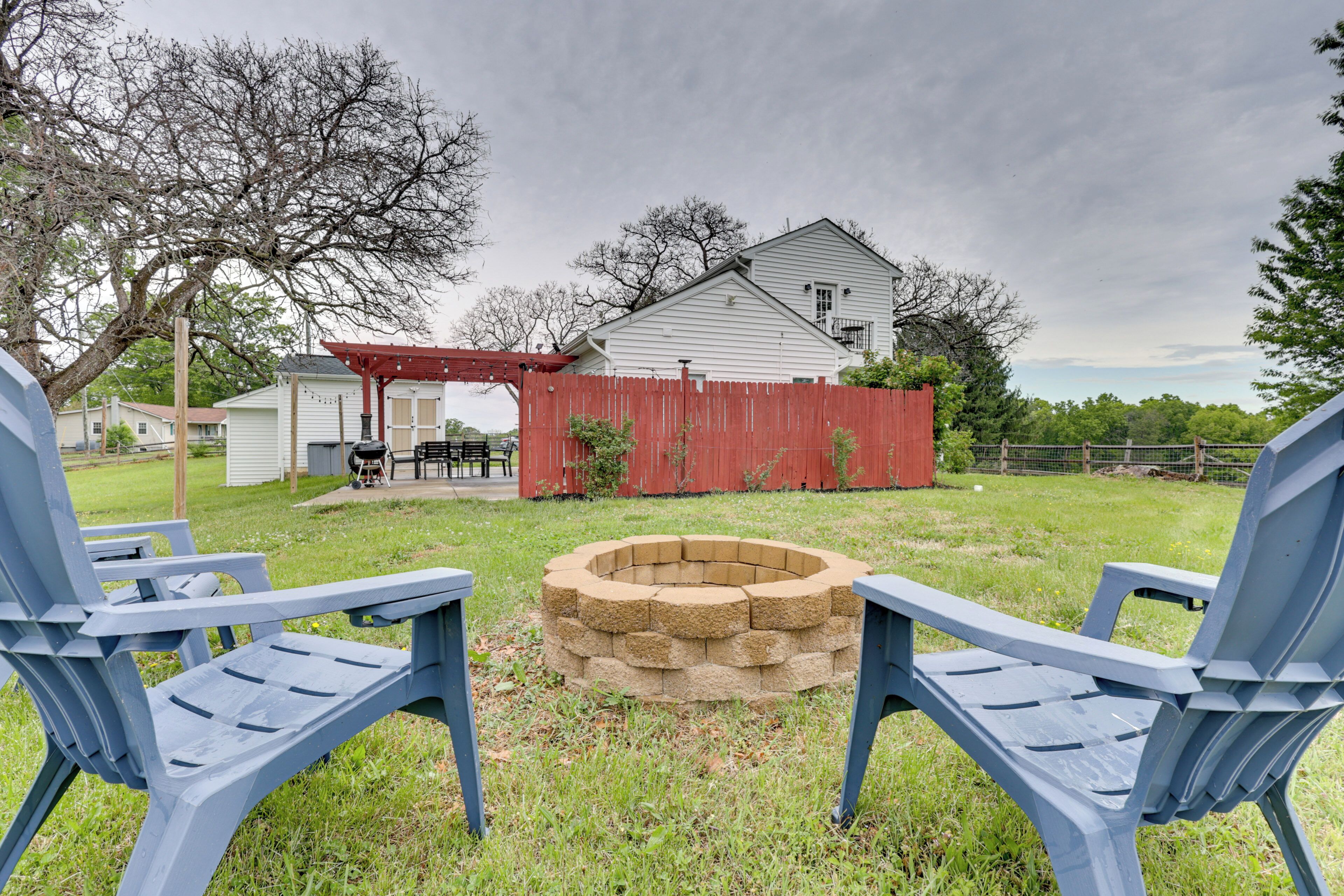 A backyard with a fire pit, two blue Adirondack chairs, a red wooden fence, and a white house with a porch.