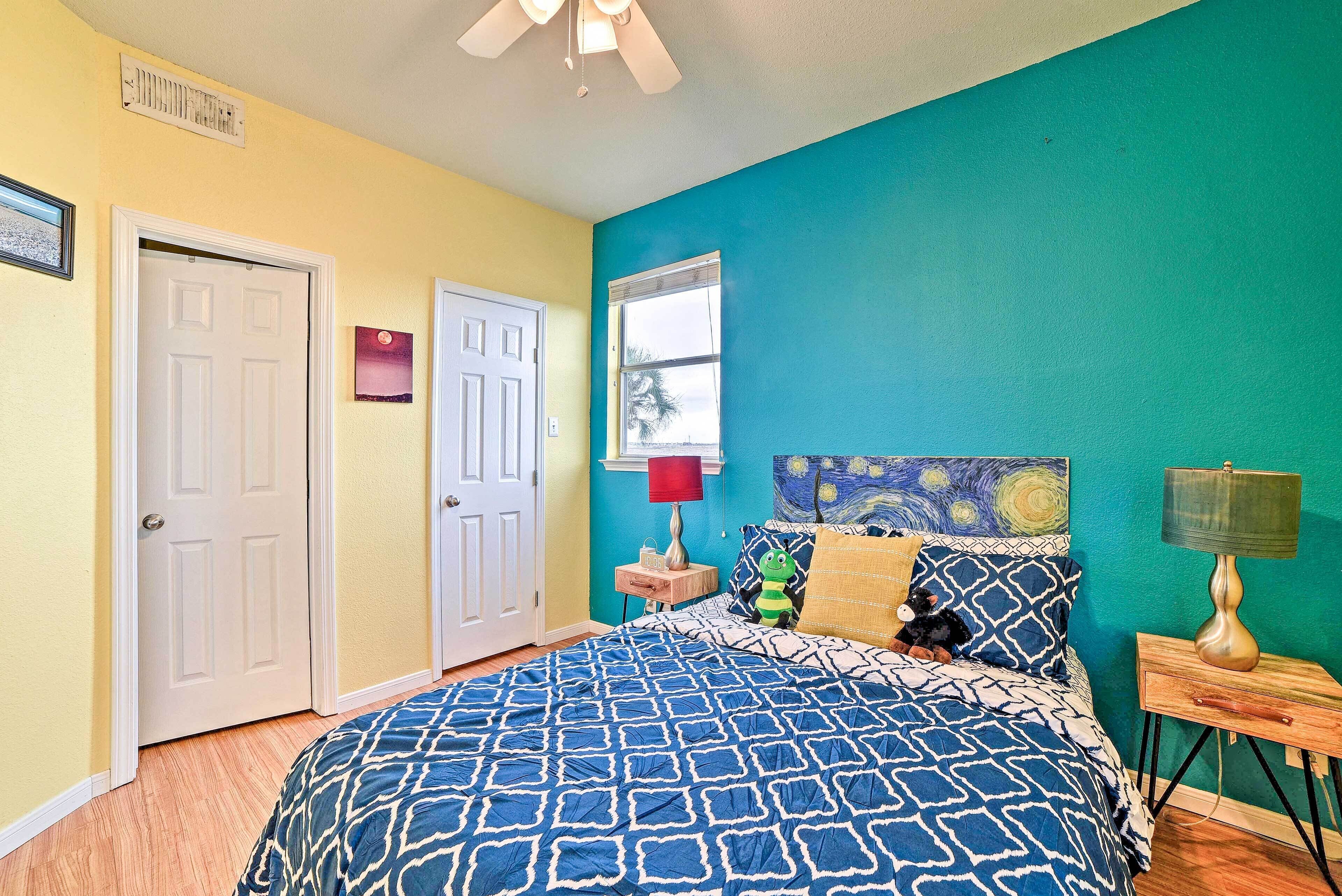 A bedroom with a blue patterned bedspread, a window with a view, a ceiling fan, and a nightstand with a lamp.