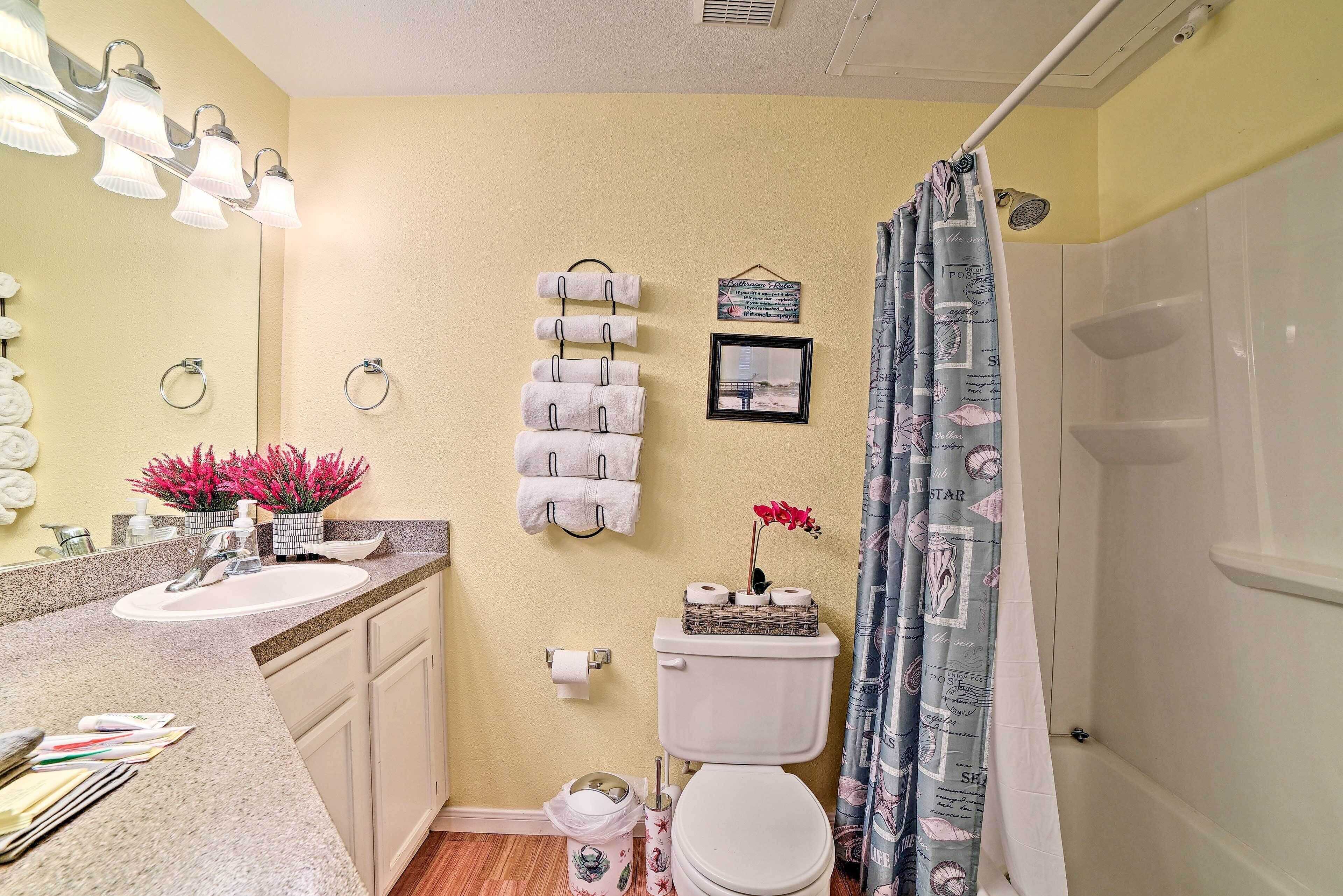A bathroom with a sink, toilet, and shower. There are towels hanging on a rack, a trash can, and a framed picture on the wall.
